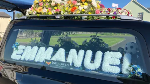 BBC A black hearse with Emmanuel's name printed in white in the window. There are colourful summer flowers on the roof of the hearse.
