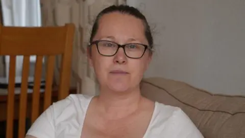 Shaun Whitmore/BBC Tracy Dolan sits on a cream sofa inside a holiday rental home. She is slightly smiling. She has dark hair that has been tied back behind her head and she wears a white T-shirt. 