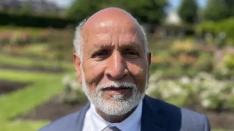 Mohammed Arshed has light brown skin with balding grey hair and a grey beard. He is wearing a grey suit and flower beds can be seen behind him