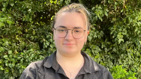Simon Dedman/BBC Alex McCormick stands in front of a green bush on a sunny day and smiles at the camera. They have brown hair that is tied up behind their head, glasses and a black shirt with a necklace around their neck. 