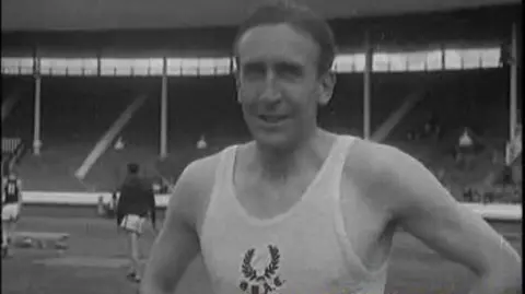 A black and white image of Alan close up to camera in a white athletic vest standing in a stadium with a couple of other athletes in the background and empty stands