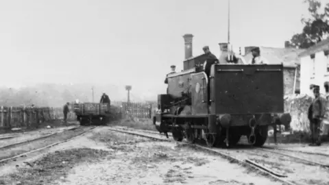 Tim Osborne's "Images of the Past" Collection An old black and white photograph of railway tracks with the railway engine arriving at the end of the line to collect a wagon at the end of it There are two figures on and near the wagon and in the foreground a man wearing a bowler hat standing in front of a house