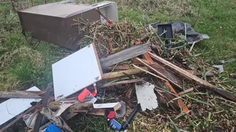 Fly-tipped wasted which has been dumped at the side of a road. There a number of plastic cans, aerosol cans, snapped wooden planks, garden items and a large wooden sideboard. 
