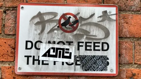 A white sign which says 'do not feed the pigeons' which has been covered with stickers and graffiti.