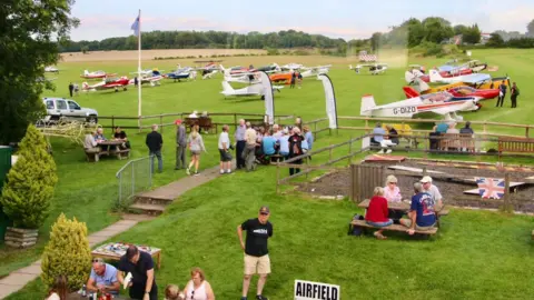 Barrie Jay Dozens of light aircraft are parked in rows on the grass airfield while, in the foreground, people gather around picnic tables in a fenced area.