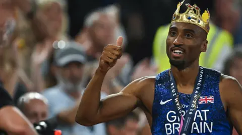 A man with a crown on his head gives a thumbs up with his right hand. He is wearing a blue top including the words Great Britain and the crowd in the background is blurred. 