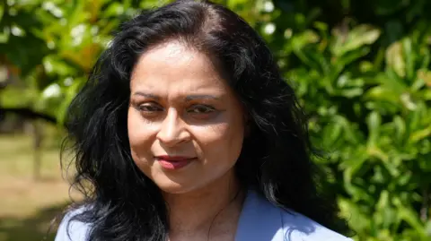 A woman with black hair wearing a blue suit smiling in front of a tree