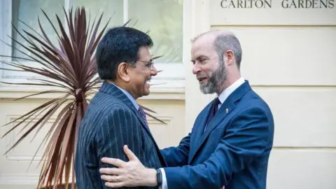 Reuters UK Business Secretary Jonathan Reynolds embraces India's Minister of Commerce and Industry Piyush Goyal when they met in London last week to finalise the trade deal. They are both wearing navy suits and smiling widely.