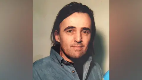 Sussex Police A man in a denim blue shirt with long black hair and a short black beard.