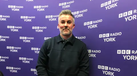 BBC/FREYA COX York and North Yorkshire's elected Labour mayor David Skaith is smiling straight at the camera. He is wearing a dark jacket and standing in front of the purple and white BBC Radio York logos.