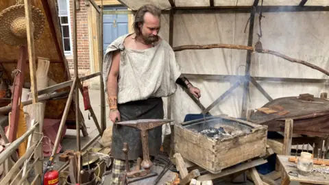 A man stokes a fire as he demonstrates how a Roman blacksmith would have worked.  He wears an old apron and a toga-style top tied at the shoulder.