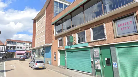 Google The picture shows the exterior of the Corn Exchange building in Exeter, viewed from Market Street on a bright day.
The building is a large, mid-20th Century brick structure with a flat roof and a mix of brickwork, concrete and glass. On the left is a taller section with long vertical rows of pale-coloured window panels running up the façade. To the right, the main frontage steps down towards the street, with several floors of windows.