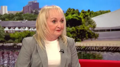 Jenni sits on a red sofa in the BBC Breakfast studio. She has shoulder-length blonde hair. She wears a white top under a grey blazer