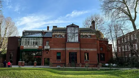 Getty Images Rear view of Leighton House in London on a clear day, showing its red-brick facade, large glass conservatory, and the distinctive golden dome of the Arab Hall, with two people walking on the lawn in the foreground and leafless trees surrounding the building.