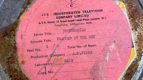 Century 21 Films  A rusty metal container with a pink label on it saying the material inside is from an episode of Thunderbirds called Trapped In The Sky. It has branding on it from 'Incorporated Television Company Limited'.