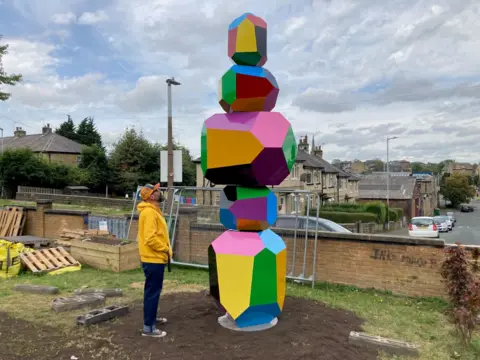 A man in a yellow hooded top and baseball cap stood looking up at a 4m multi-coloured sculpture with five separate multi-sided sections.