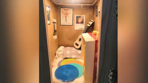 The Rumble Museum The locker with a record player toy. There are posters on the wall for music groups such as Nirvana. There is a guitar on the bed and record players beside it.