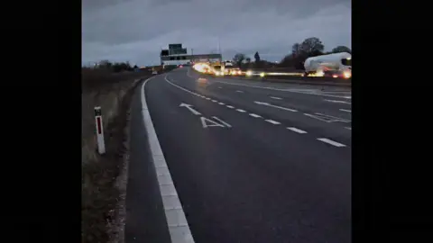 Faith Best The A14, a three-lane road, without traffic on it on one side. In the foreground is a roadmarking indicating the A1 while in the background there is signage over the road.