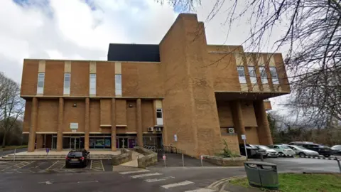 Google Peterborough's Regional Pool - a brutalist structure in an abstract shape, with small windows across the top level. In front of it are a number of parked cars.