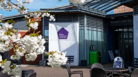 The glass front of a university building. A purple and white sign outside reads Bishop Grosseteste University and the picture has been taken behind a tree with white blossom in shot.