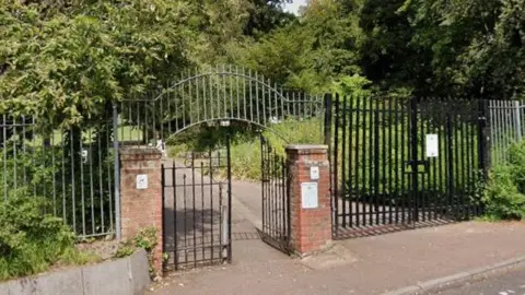 Google The entrance to Belle Vue Park - a black metal gate for people is flanked by brick pillars. A larger metal gate for vehicle access is next to it. Beyond the fencing are trees and bushes and a path.