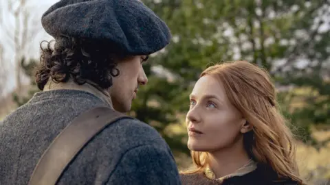 Starz Jamie Roy as Brian Fraser and Harriet Slater as Ellen MacKenzie in 18th Century dress stand in a field looking at each other with trees behind them. Brian wears a dark hat and Ellen has long ginger hair.