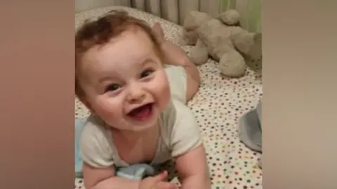 Baby Preston Davey smiles at the camera dressed in a white vest. He appears to be lying in a cot with a star-covered bedsheet.