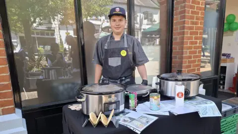 Dexter stood in his all grey chef uniform stood in front of slow cookers on a table with recipe cards, spices, tins and flatbreads on the table.