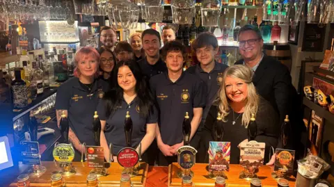 Camra A number of staff are behind the bar in their uniform next to the owners. 