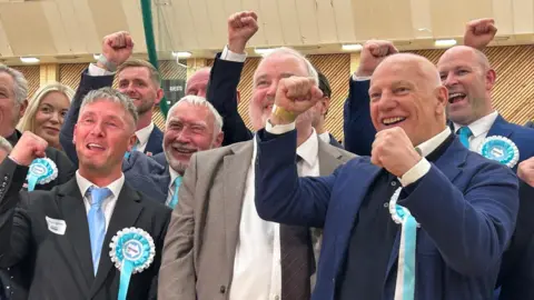 A group of male Reform representatives have their fists aloft in celebration. They are smiling as if they are mid-cheer and are wearing suits and turquoise rosettes. There is one woman at the back of the group on the left.