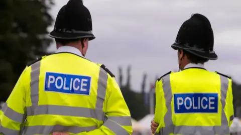 Two male West Midlands Police officers stand with their backs to the camera. They have helmets on and fluorescent jackets with "Police" on the back. Trees can be seen in the background.
