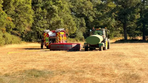 Brian Seeger A tractor with a water bowser in a field next to a fire engine which has a yellow hose laid on the ground. The field is very dry and there are woods in the background.