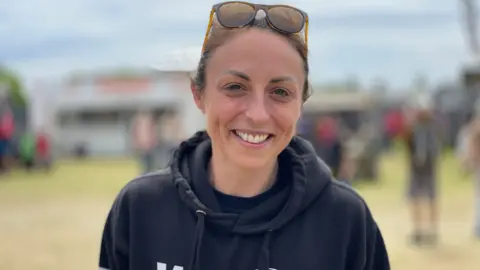 Hayley Bell has dark hair tied back with sunglasses resting on her head. She is smiling and wearing a black hooded jumper. She is standing outside with groups of people behind her.