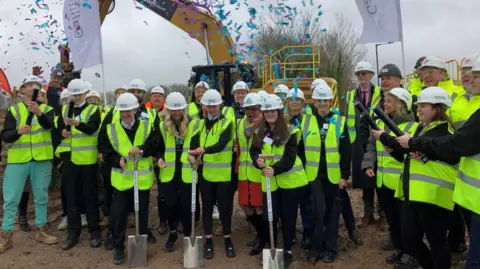 A large group of people, many of them teenage children, stand together wearing white hard hats and high-vis jackets at the site of the new youth centre for Knowle West in Bristol. Three of the children at the front are holding spades and England and Bristol rugby player Ellis Genge is visible in the group