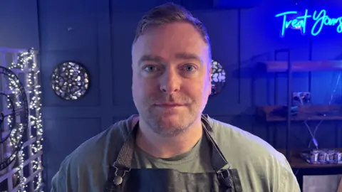 Daniel is looking directly at the camera. He is wearing a green t-shirt and has a black apron on top. He is standing in front of a grey panelled wall and there is a blue neon sign to the right of the image which says 'treat yourself'. 