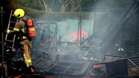 A firefighter stood in a badly fire-damaged garden. The photo shows the ruins of what could be a shed, as well as a charred wheelbarrow and a fire-damaged children's play house.