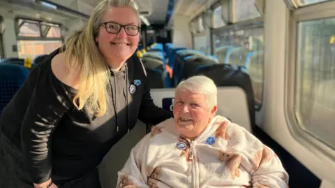 Ellie McWilliam is standing in the train aisle beside her mum Jean who is in a wheelchair. Both are smiling to the camera. Ellie is wearing black and Jean has a warm fleece with teddy bears on it, she is wearing sheepskin mittens. 