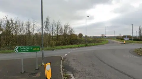 Rural roundabout, with a green sign indicating "Peterborough, Oundle" to the left alongside a yellow bollard. A white road sign is visible in the background.