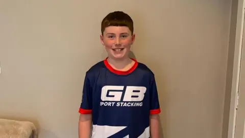 Joanna McStravick Olly is a boy with brown hair and is wearing a blue sports top with "GB sport stacking" written on it in white text. He is standing in front of a wall in a living in front of a cream-coloured wall.