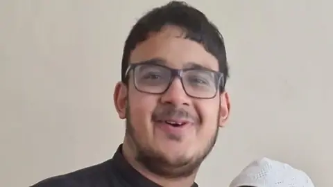 A young man with black hair, square black glasses, a beard around the edge of his jaw and a moustache. He is smiling and looks as if he might be midway through speaking. He is wearing a black shirt with a small collar.