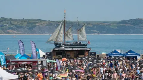 Scoop The festival is set up at Weymouth Peninsula. Crowds can be seen gathered around stalls and there is a ship with white sails in the sea in the background, with green hillside seen in the distance.