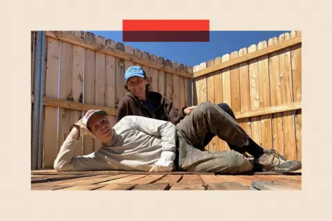 A man and a woman pose on some decking. The man is lying down on the floor, and the woman is sitting behind him. They are both wearing baseball caps and similing.