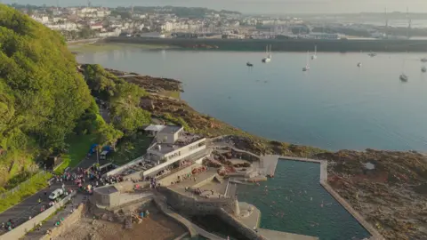 Ross Yeates A drone photo showing the walkers gathering at the bathing pool. There are many swimmers in the pool. There are yachts moored in the water beyond and the town can be seen on the far shore.