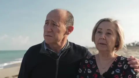 Luis Elena Blas and Roser Vento Sancho stand on a beach. He has her arm around her as they look out to sea. Mr Elena Blas is balding, with short, greying hair, and wears a dark, zipped jacket and a blue shirt. Ms Vento Sancho has short, fair hair and wears a dark, patterned dress.