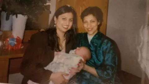 Two women smile at the camera and hold a baby wrapped in a white blanket