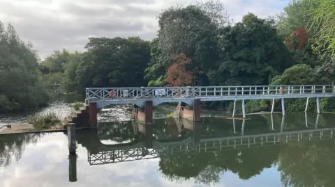 Robin A weir with a walkway over it is reflected in the water. Behind it are trees and the river heading downstream