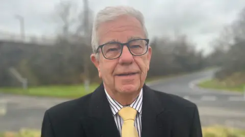 A man with grey hair and black glasses looking straight at the camera with a road behind him, wearing a black overcoat a yellow tie and a striped shirt