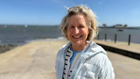 Heather Gilmore has blonde hair and is smiling. She is wearing a pale blue puffer coat with a stripped cardigan below and blue t-shirt. In the background there is the lough and the slipway down to it. 