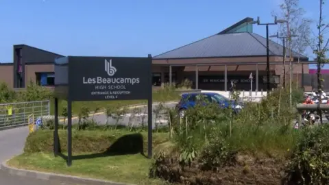 BBC Les Beaucamps High School has been photographed showing a dark grey sign that reads Les Beaucamps High School. Behind the sign is the school with a light brown exterior and a light grey roof. 