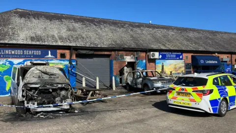 A police car is parked outside the scene, which has been cordoned off with police tape. The two destroyed vehicles outside the shop are blackened through fire-damage. The wall has been damaged around the entrance to the building.